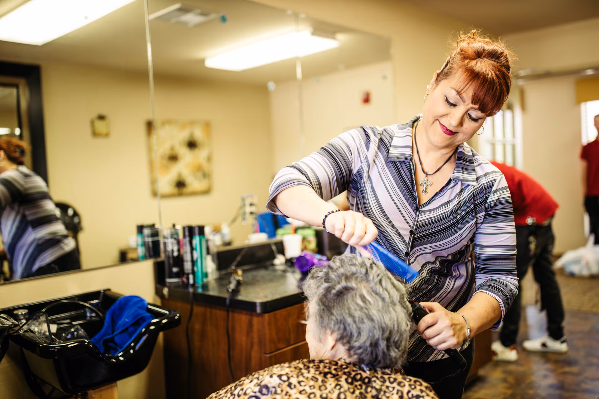 A hairstylist combs the hair of an elderly woman seated in a salon chair inside a hair salon. The hairstylist is smiling and wearing a striped shirt, while the elderly woman has short gray hair and is wearing a leopard print top. The salon has a large mirror, various hair products on the counter, and a black sink with a blue towel.