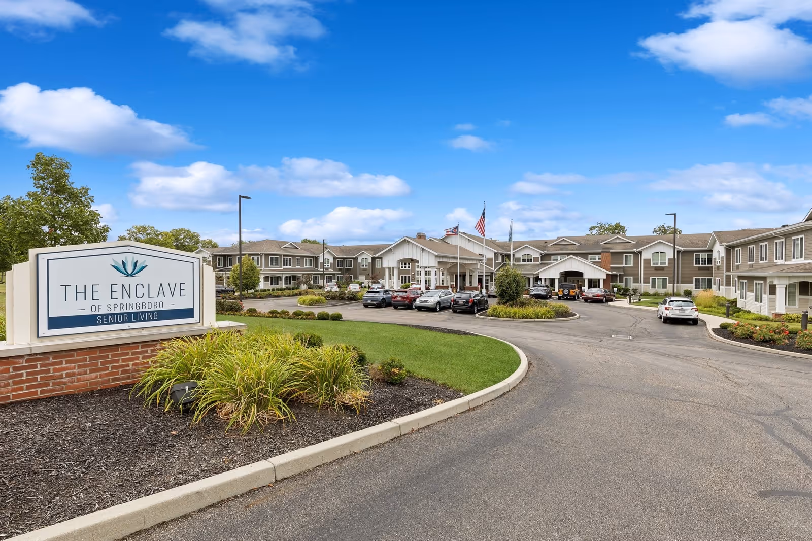 Exterior view of The Enclave of Springboro senior living facility showing a large driveway with parked cars, landscaped greenery, and a clear blue sky with some clouds.