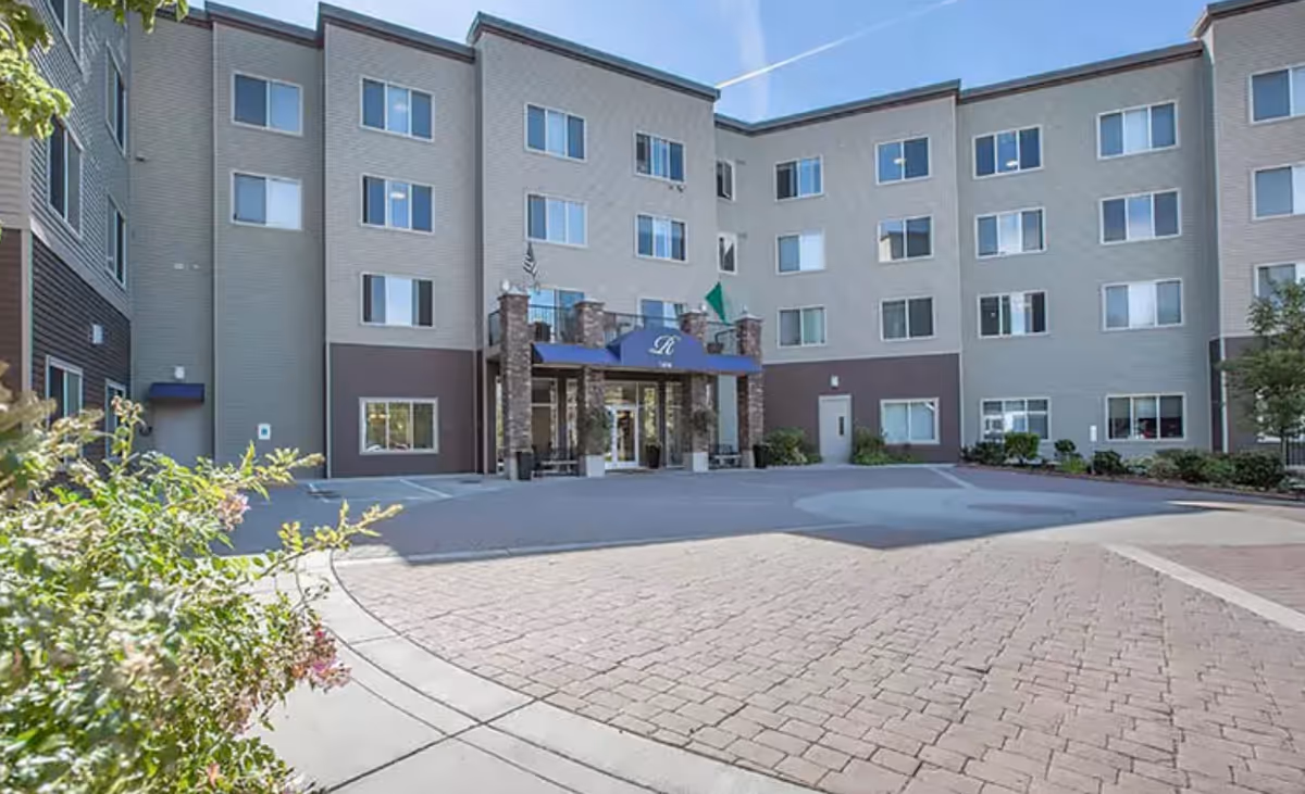 Front entrance of a multi-story senior living building with a covered porte-cochère and a paved circular driveway.