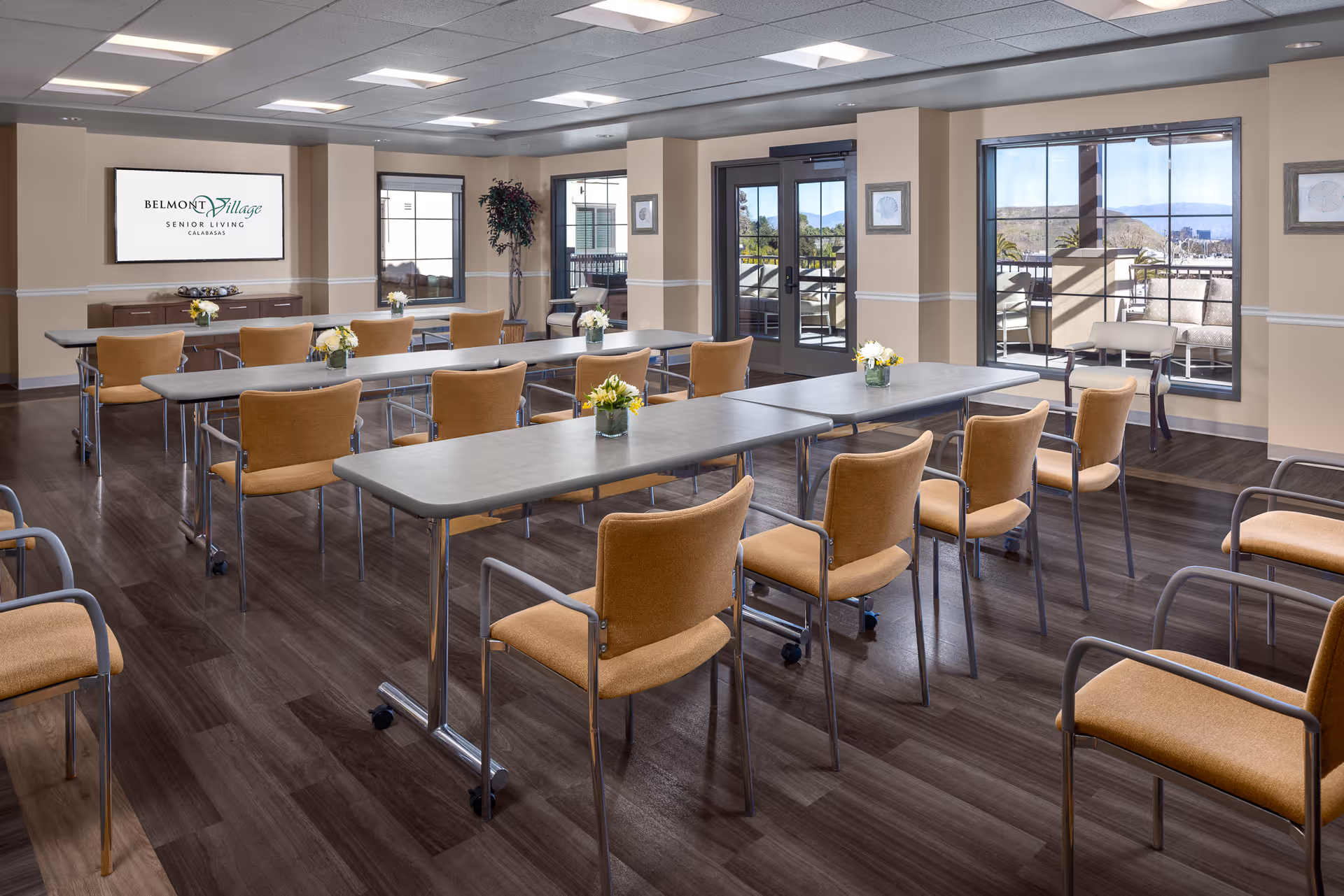 A bright and spacious meeting or activity room in Belmont Village Senior Living Calabasas with several tables arranged in rows, surrounded by cushioned chairs with armrests. The room has large windows and glass doors that provide a view of an outdoor patio area with seating and scenic mountains in the background. The walls are beige with white trim, and there is a large screen on one wall displaying the Belmont Village Senior Living Calabasas logo. Small flower arrangements are placed on the tables.