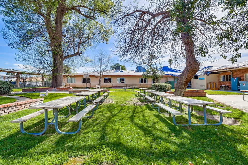 Outdoor area of Colonial Gardens Nursing Home featuring several wooden picnic tables with metal legs arranged on a grassy lawn. Large trees provide partial shade, and a single-story building with windows and a basketball hoop is visible in the background under a partly cloudy sky.