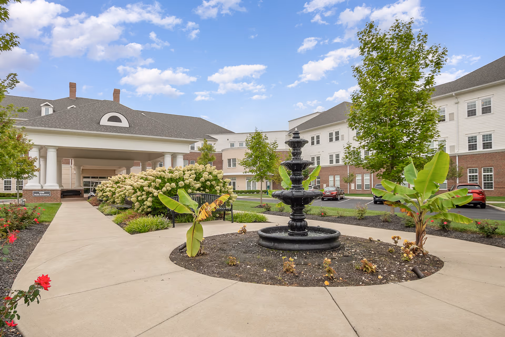 Outdoor view of Wesley Woods facility showing a circular walkway with a black tiered fountain in the center, surrounded by small plants and trees. The building has white siding with brick accents and multiple windows under a partly cloudy sky.
