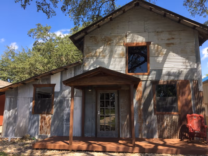 Exterior view of a rustic building with corrugated metal siding and wooden trim. The building has a small covered porch with two wooden posts and a glass door. There are three windows with wooden frames and a red chair on the right side of the porch. Trees and a blue sky with some clouds are visible in the background.