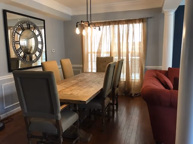Dining room with a wooden table and upholstered chairs, a large round wall mirror, hanging light fixture, sheer curtains, and a red sofa.