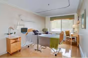 A bright and clean patient room with a hospital bed, medical equipment, a wooden nightstand with a potted orchid, a desk with a chair near a window with blinds, and a lamp on the desk.