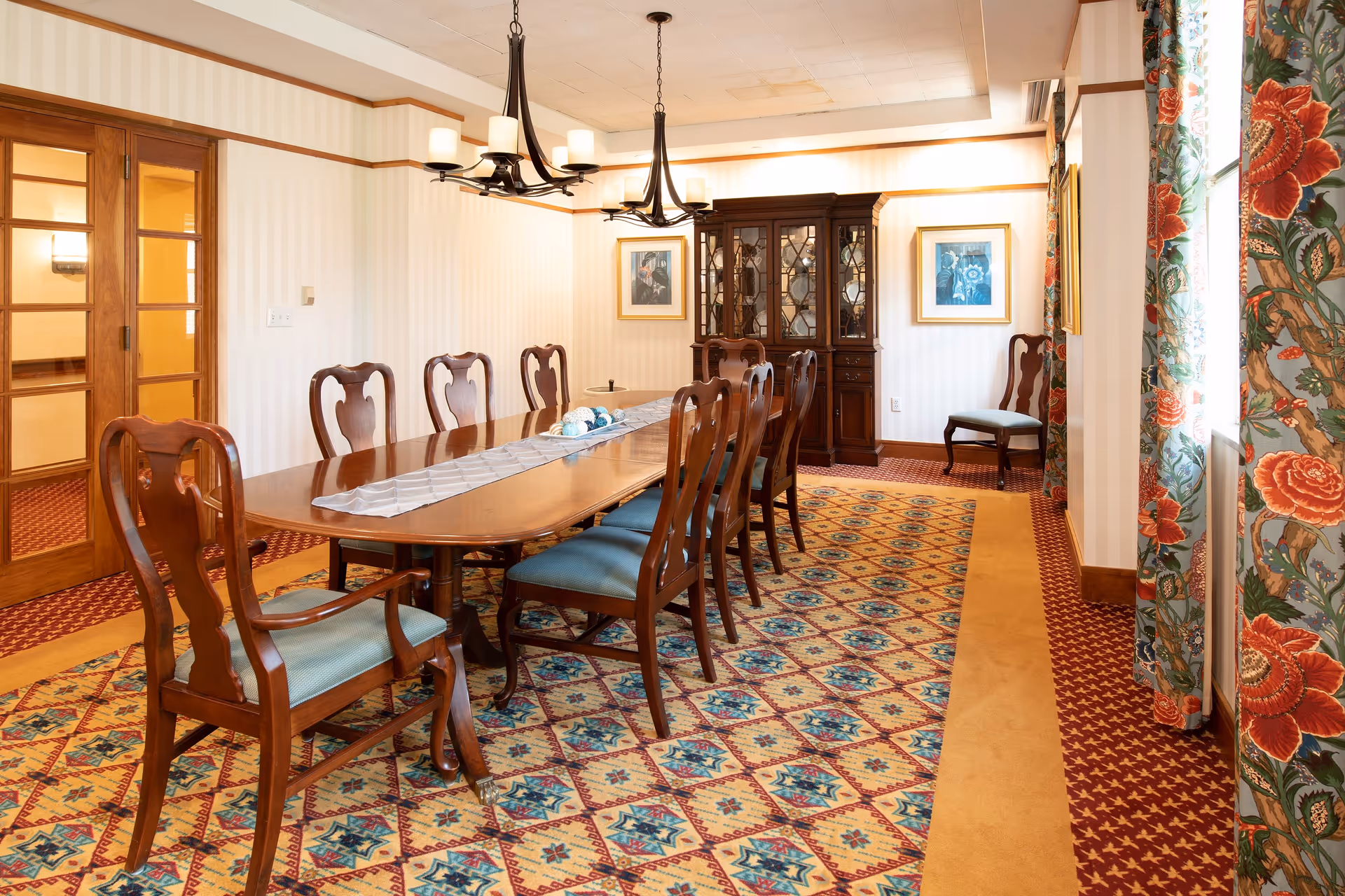 A spacious dining room featuring a long wooden table surrounded by elegant chairs, with decorative curtains and a patterned carpet. A china cabinet is visible in the background, along with framed artwork on the walls.