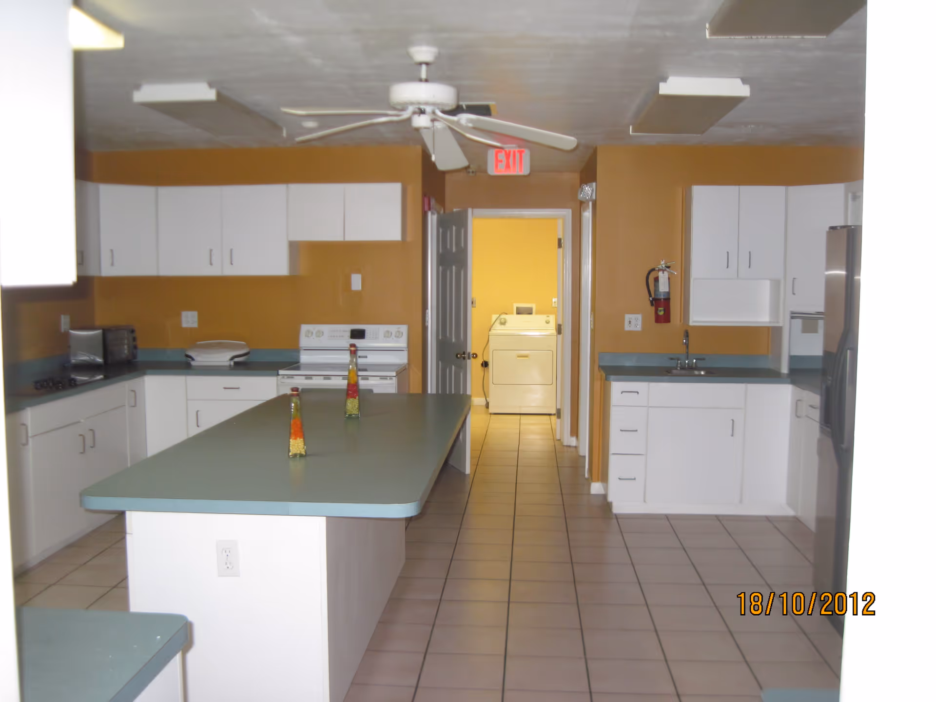 Spacious communal kitchen with a central island, white cabinets, teal countertops, and a view into a laundry room.