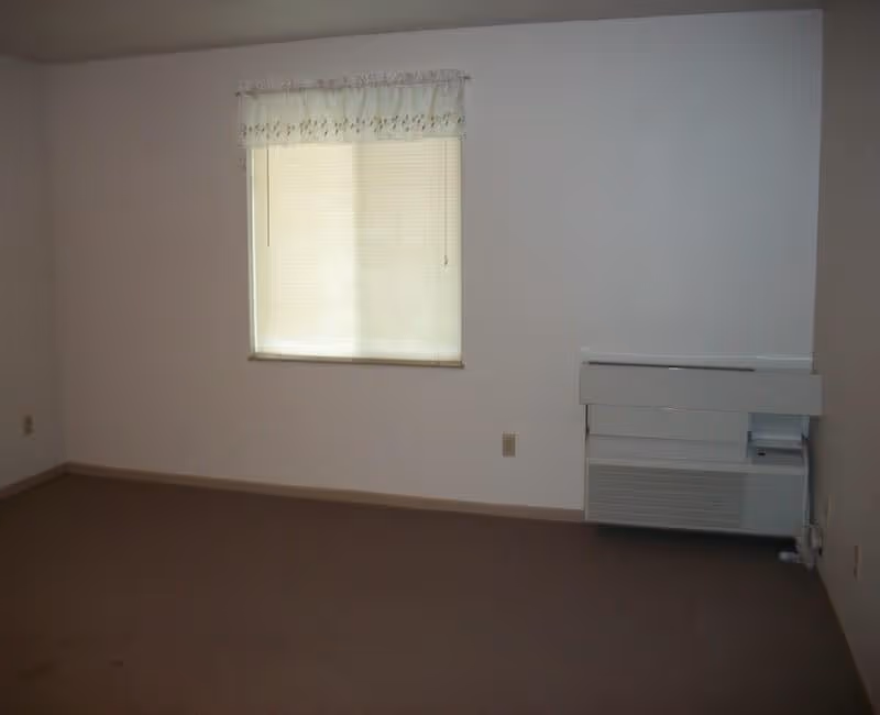 Empty room with beige carpet, white walls, a window with closed blinds and a floral valance, and a wall-mounted heating or air conditioning unit below the window.