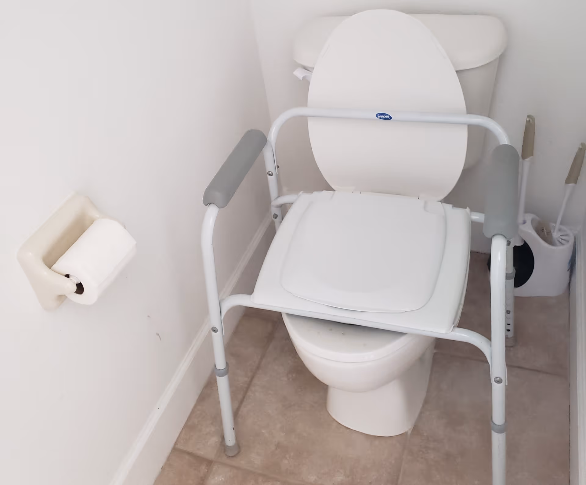 A white toilet with an open lid in a bathroom, equipped with a white and gray raised toilet seat frame with armrests for support. To the left, there is a toilet paper holder mounted on the wall with a roll of toilet paper. In the corner to the right, there is a toilet brush and plunger set on the tiled floor.