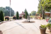 Outdoor patio area with a concrete walkway, a small gazebo, a water fountain, potted plants, and a wooden swing set surrounded by trees and bushes under a clear sky.