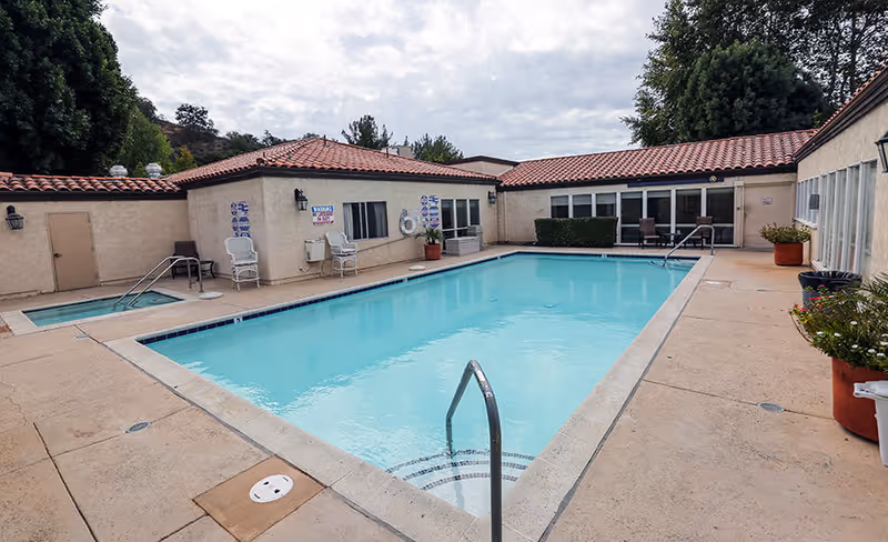 Outdoor swimming pool with clear blue water surrounded by beige concrete deck and single-story buildings with red tile roofs. There are chairs and potted plants around the pool area, and a smaller hot tub is visible to the left. Trees and cloudy sky are in the background.