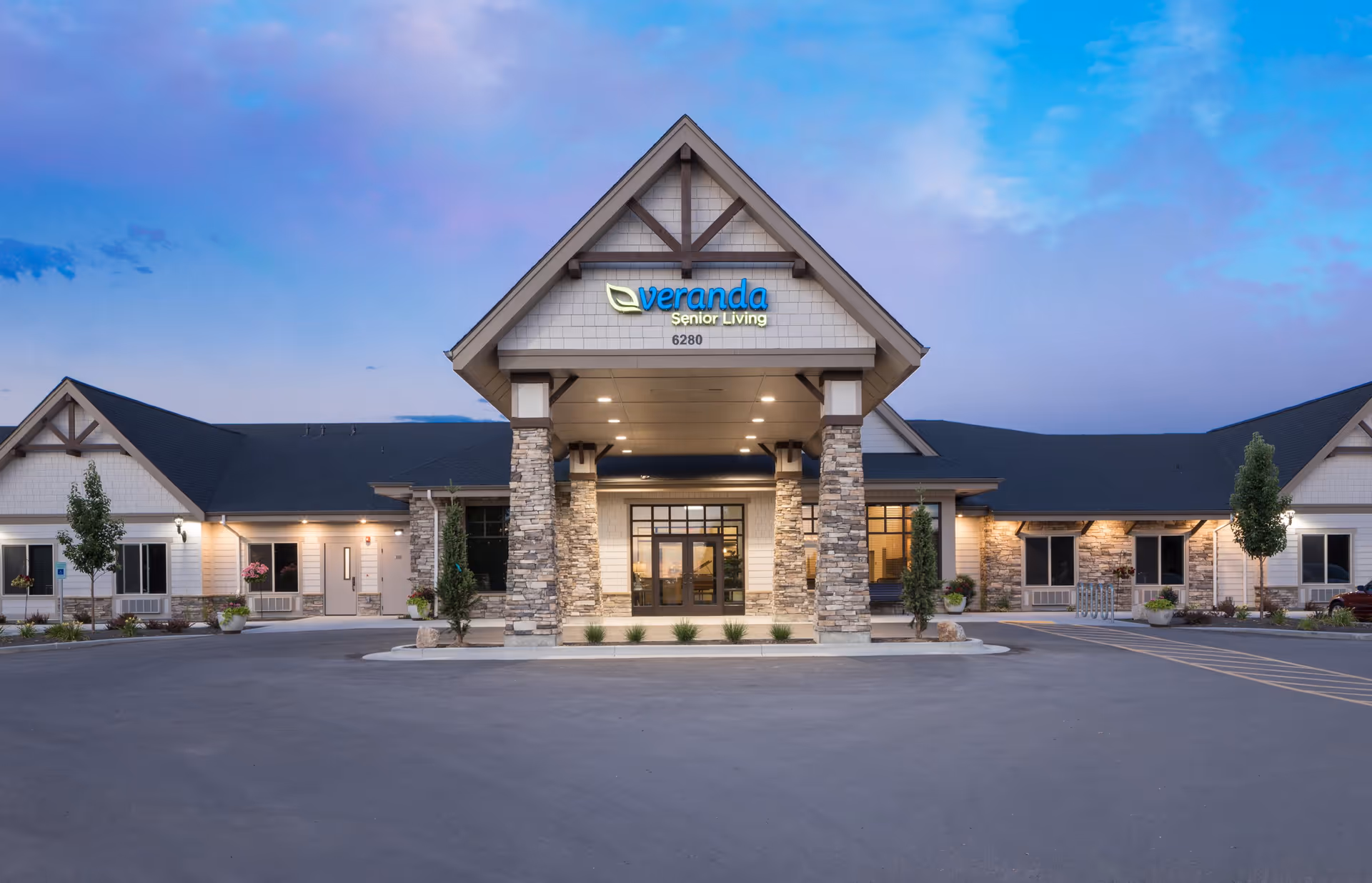 Exterior front view of a senior living facility building with stone pillars supporting a covered entrance. The building has a pitched roof and is illuminated with exterior lights at dusk. The sign above the entrance reads 'veranda Senior Living 6280'. There are small trees and plants around the building and a large paved driveway in front.