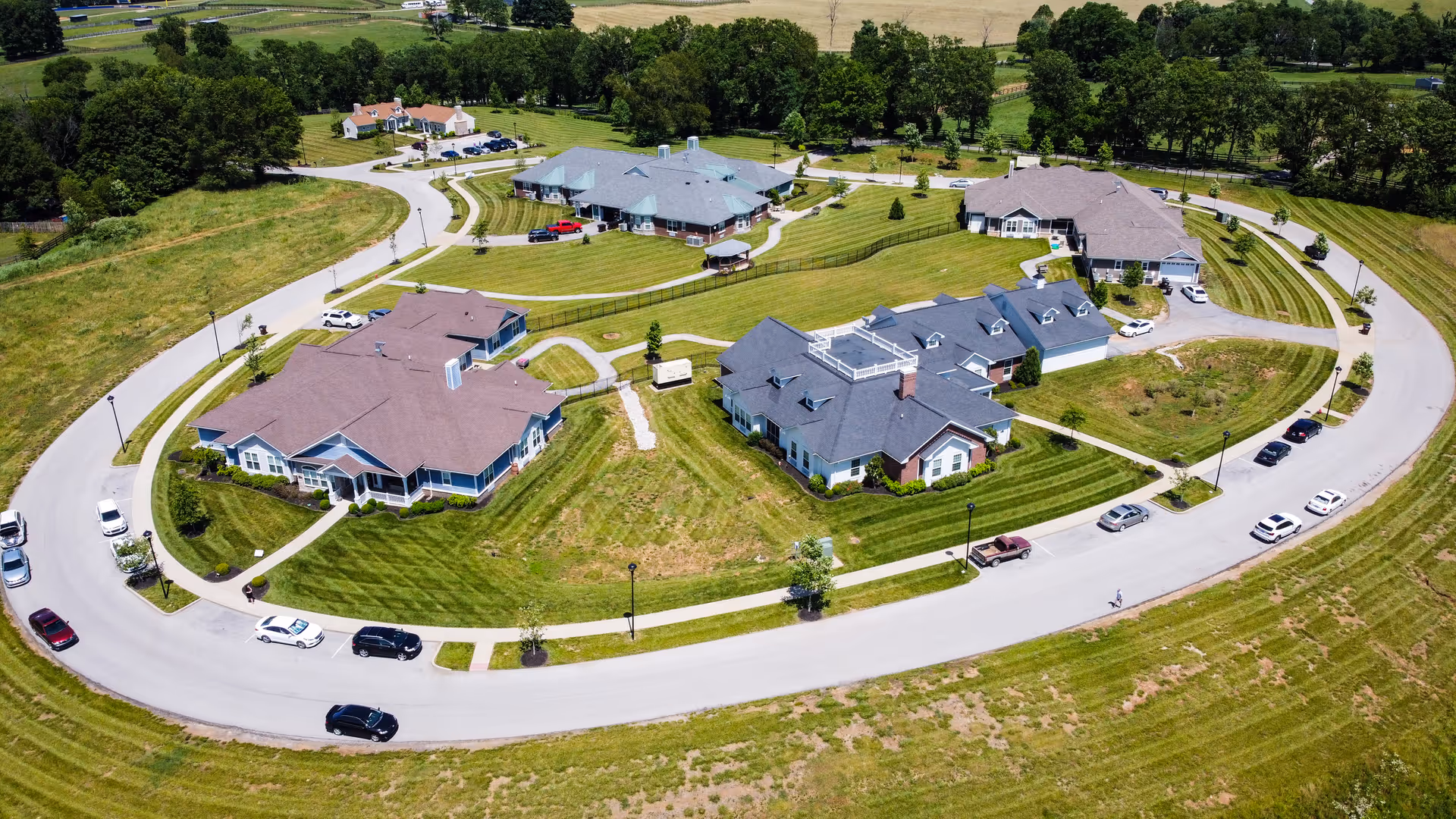Aerial view of The Homeplace at Midway senior living facility showing multiple single-story buildings with pitched roofs arranged in a circular layout surrounded by well-maintained lawns and a curved road with several parked cars. Trees and open fields are visible in the background.