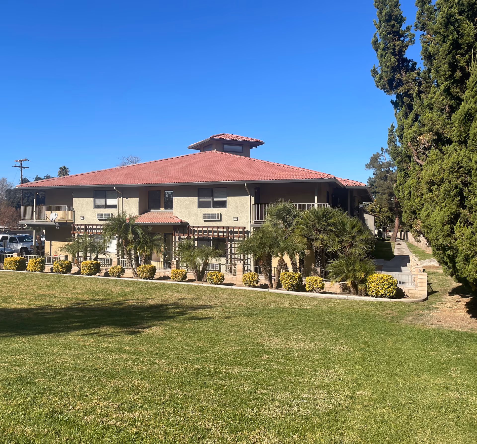 Two-story light-colored building with a red tiled roof, balconies, palm trees and a manicured grassy lawn under a clear blue sky.