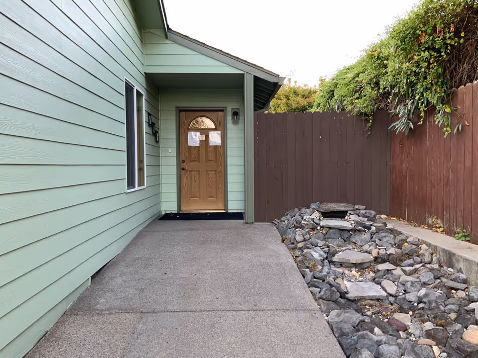 Front entrance of a pale green house with a wooden door, concrete walkway, and rock landscaping beside a brown fence.