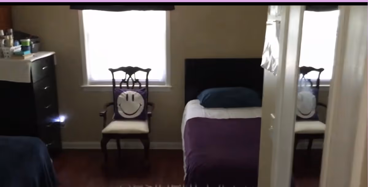Small furnished bedroom with a twin bed covered in a purple blanket, two wooden chairs with smiley-face pillows, a dresser, and a mirrored closet reflecting the room.