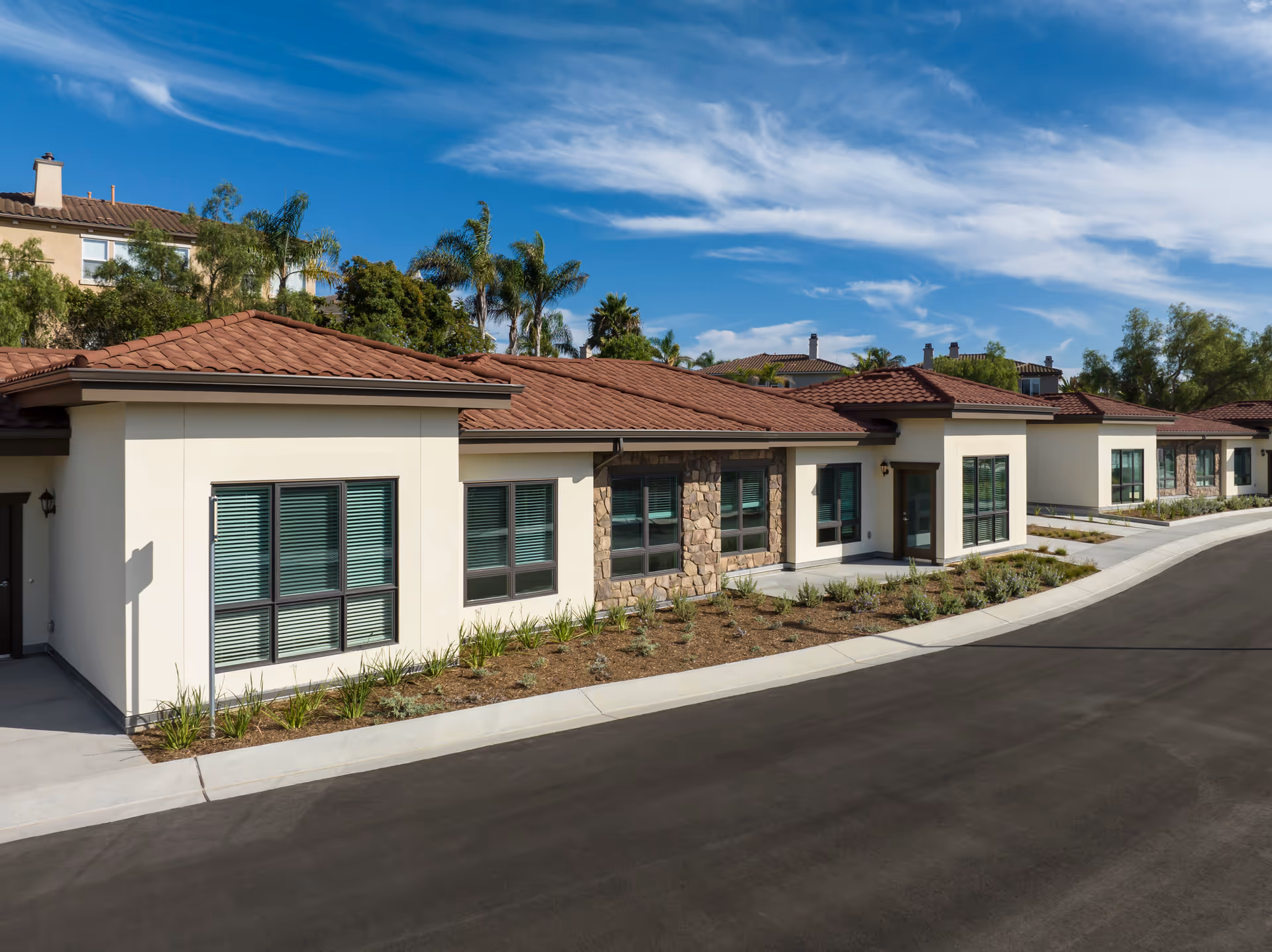 Single-story Mediterranean-style senior living building with red tile roof, stone and stucco façade, windows and landscaping along a paved driveway under a blue sky.