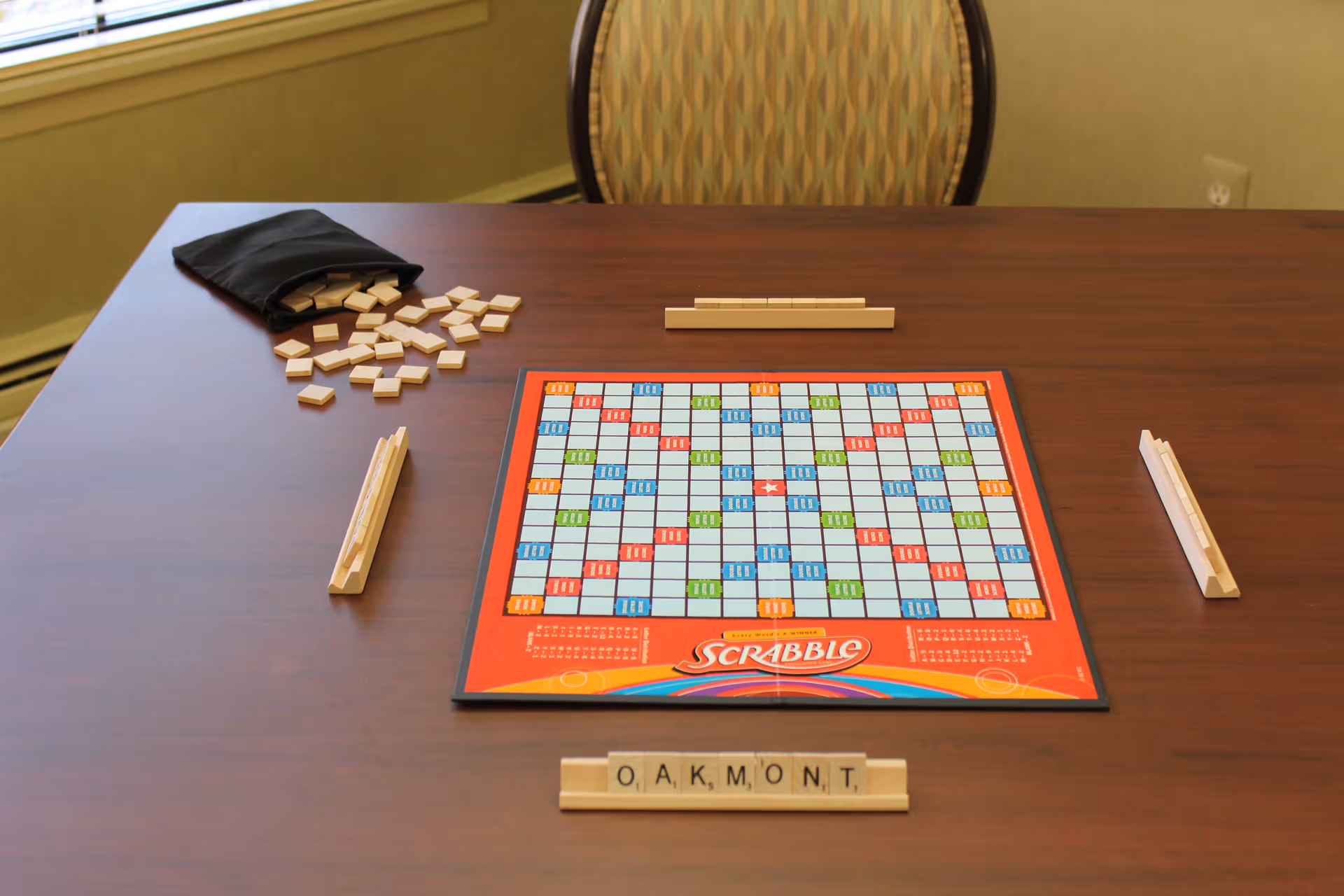 A Scrabble board game set up on a wooden table with letter tiles spelling 'OAKMONT' on a tile rack. Additional tile racks and a black bag with scattered tiles are also visible. A patterned chair is positioned behind the table near a window.