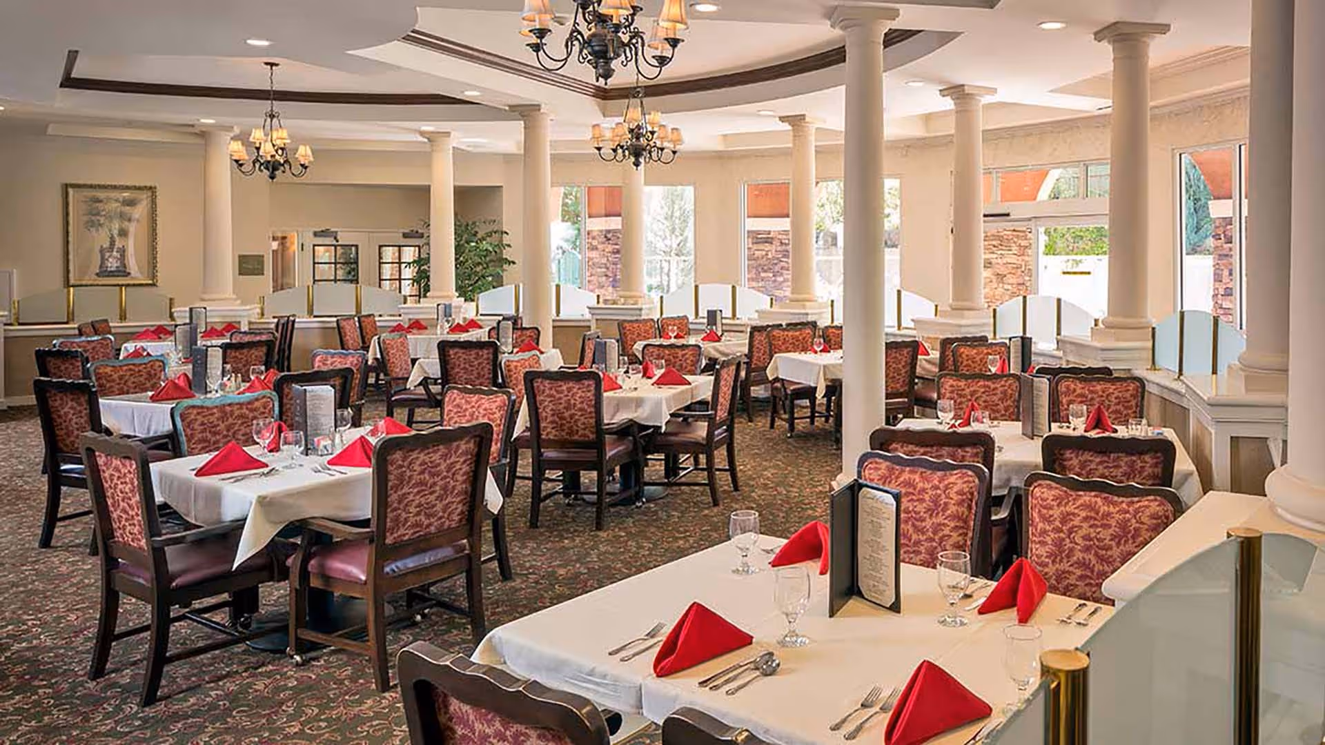 A spacious dining room with multiple tables covered in white tablecloths, each set with red folded napkins, glassware, and menus. The room features elegant chandeliers, large windows allowing natural light, decorative columns, and patterned carpet flooring.