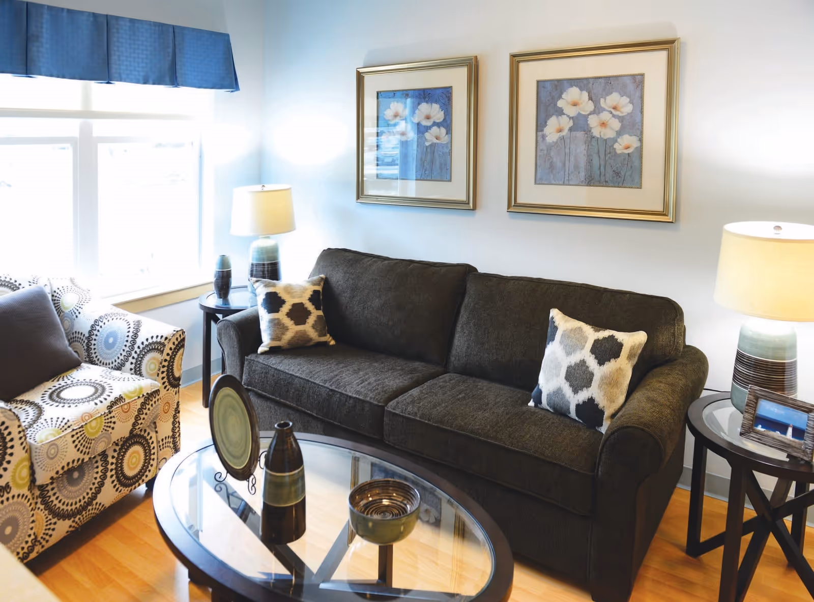 A cozy living room with a dark gray sofa adorned with two patterned pillows, a patterned armchair with a gray cushion, two side tables each with a lamp, and a glass coffee table holding decorative items. Two framed floral paintings hang on the wall above the sofa, and a window with a blue valance lets in natural light.