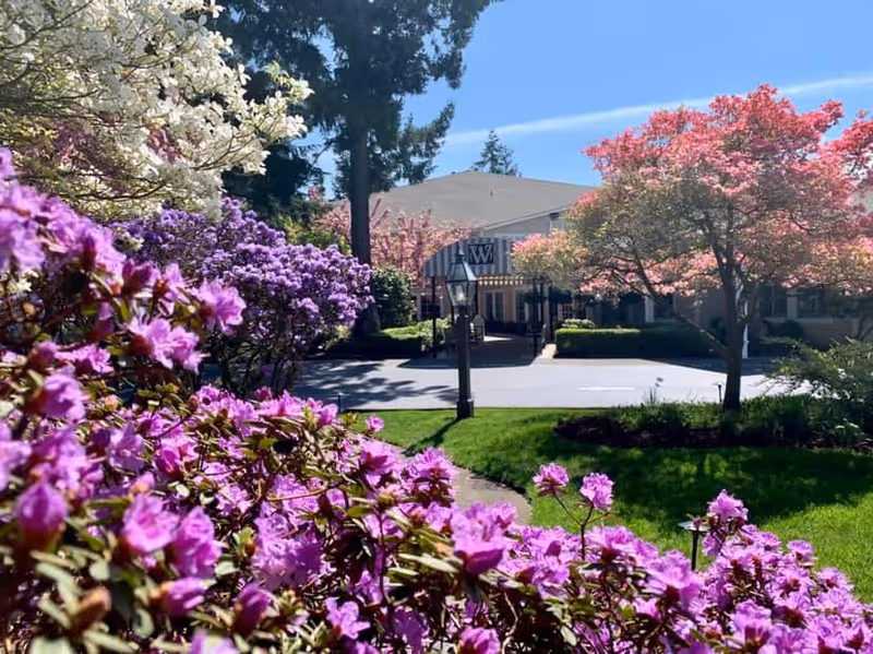 View of the Weatherly Inn Tacoma facility entrance surrounded by vibrant blooming flowers and trees under a clear blue sky.
