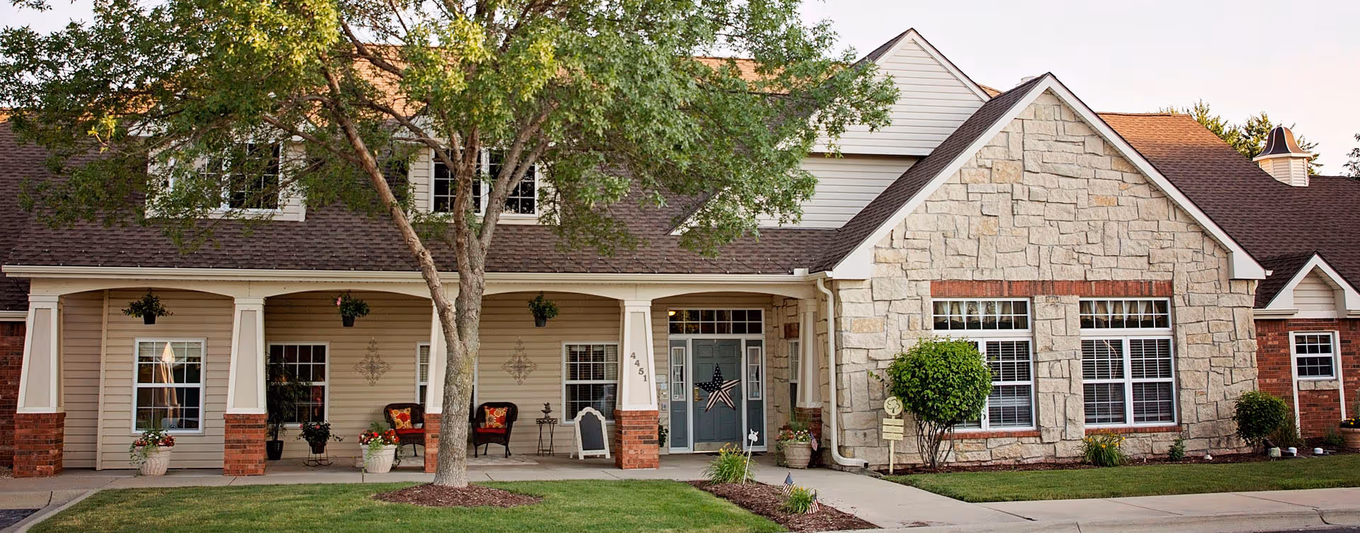 Front exterior of a single-story building with a covered porch, chairs, columns, stone and siding facade, and a landscaped lawn.