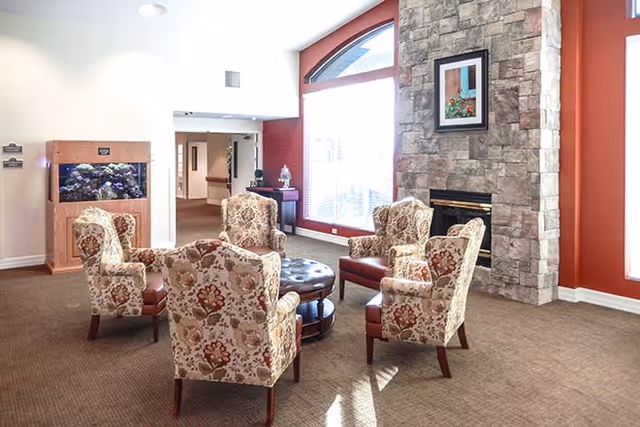 A cozy seating area in a senior living facility with five floral upholstered armchairs arranged around a round leather ottoman. The room features a stone fireplace with a framed picture above it, large windows letting in natural light, and a wooden fish tank on the left side.