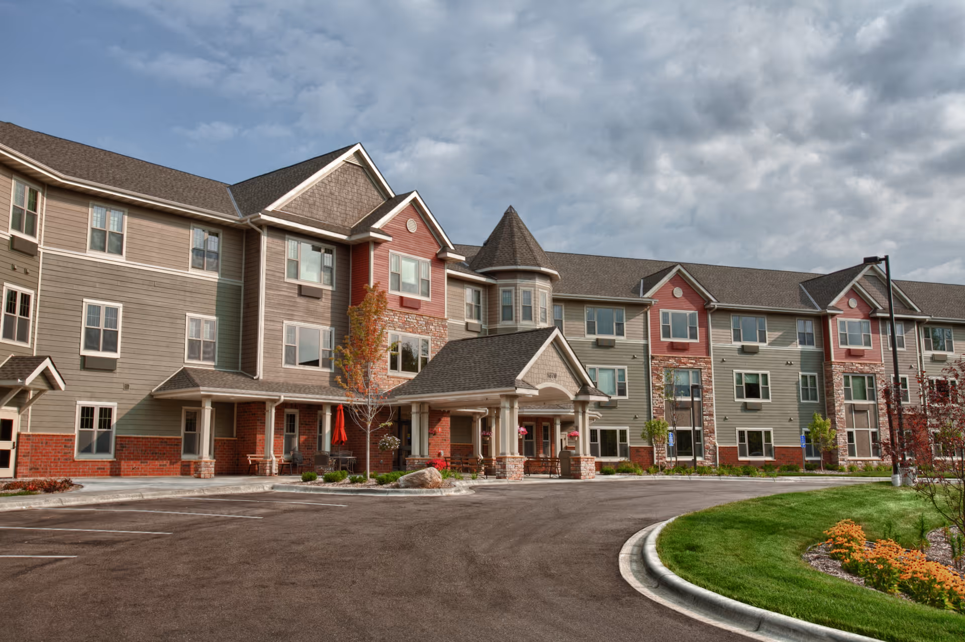 Three-story senior living building with a covered main entrance, driveway and landscaped lawn under a cloudy sky.