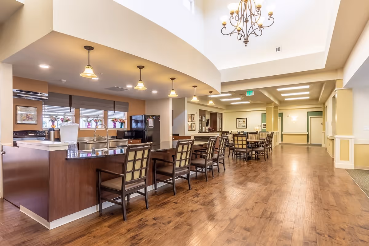 A spacious and well-lit dining area in Willowbrook Place featuring a long kitchen island with several chairs, pendant lights hanging above, and a chandelier. The kitchen area includes a refrigerator, stove, and sink with windows adorned with flower pots. The dining area has multiple tables and chairs arranged neatly on a wooden floor.