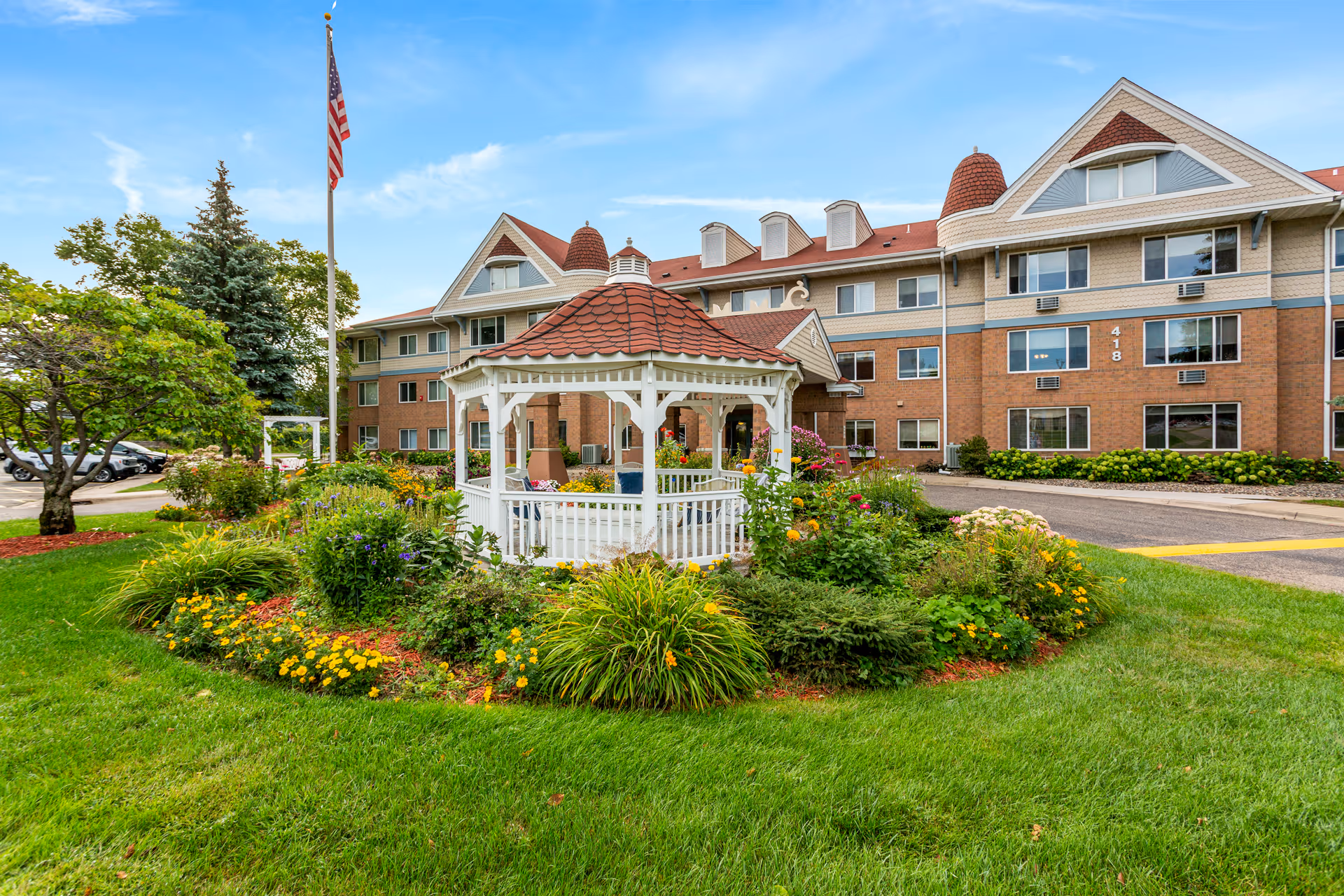 A white gazebo surrounded by colorful flowerbeds and lawn sits in front of a multi-story brick senior living building with an American flag.
