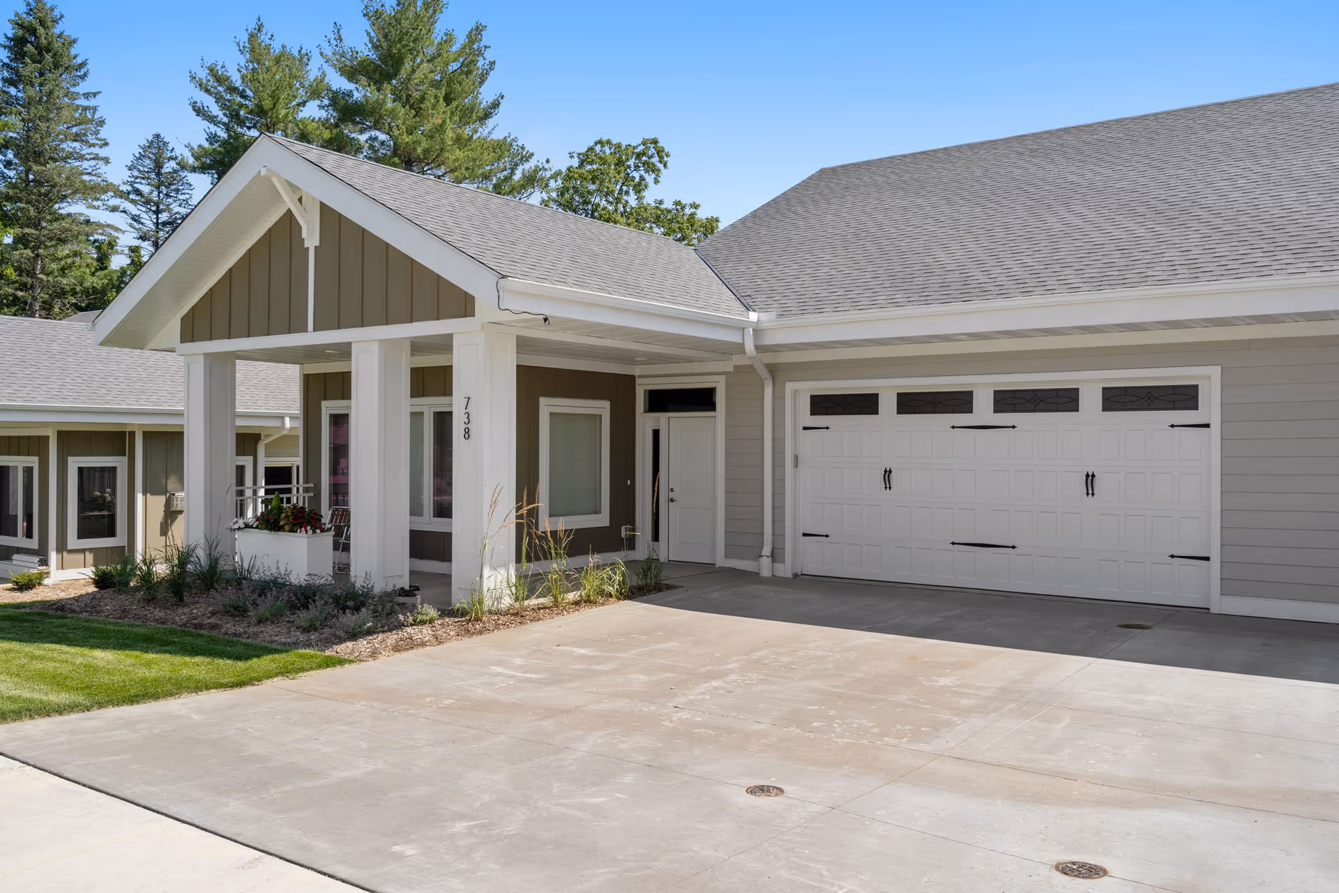 Exterior view of a residential building with a covered entrance supported by white pillars, a large white garage door, and beige siding. There are some plants and greenery near the entrance, and trees are visible in the background under a clear blue sky.