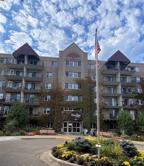 Front exterior view of Vernon Terrace of Edina, a multi-story senior living facility with balconies, ivy growing on the facade, a flagpole with an American flag, and landscaped flower beds in front.