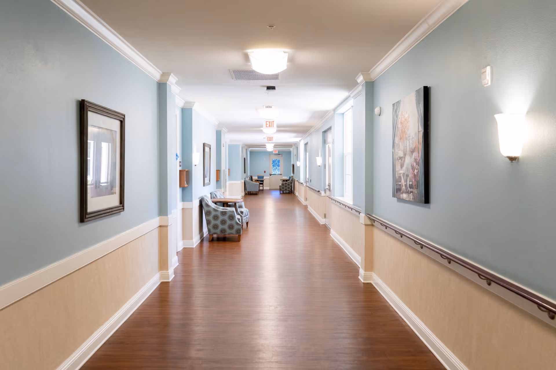 A long, well-lit hallway in a senior living facility with light blue and beige walls, wooden flooring, handrails on both sides, framed artwork on the walls, and several armchairs and small tables placed along the corridor.