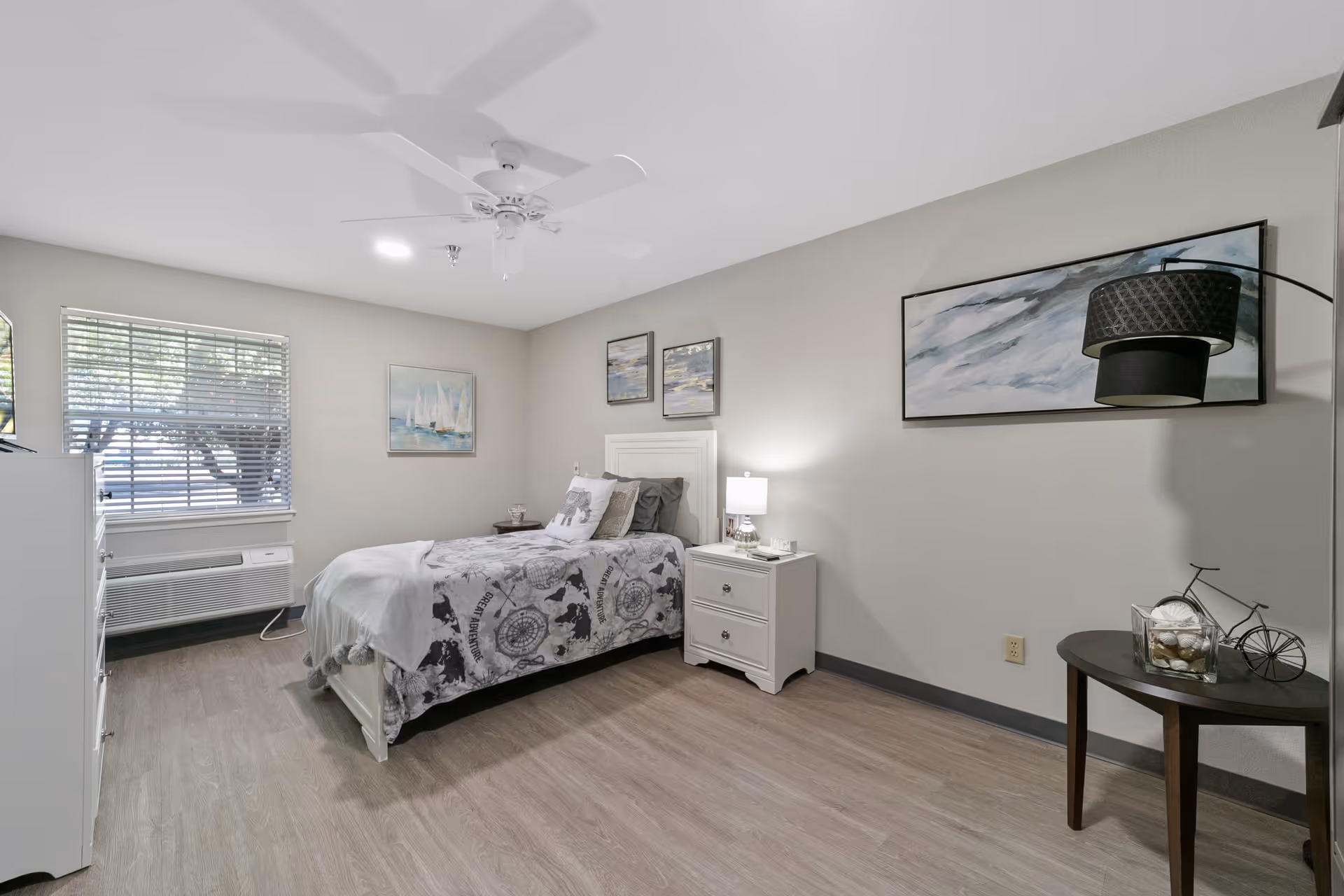 A neatly arranged bedroom with a single bed covered in a patterned gray and white blanket. The room has light gray walls, a ceiling fan, and wood flooring. There is a white nightstand with a lamp next to the bed, a small round table with decorative items, and three framed paintings on the walls. A window with blinds and an air conditioning unit is visible on the left side.