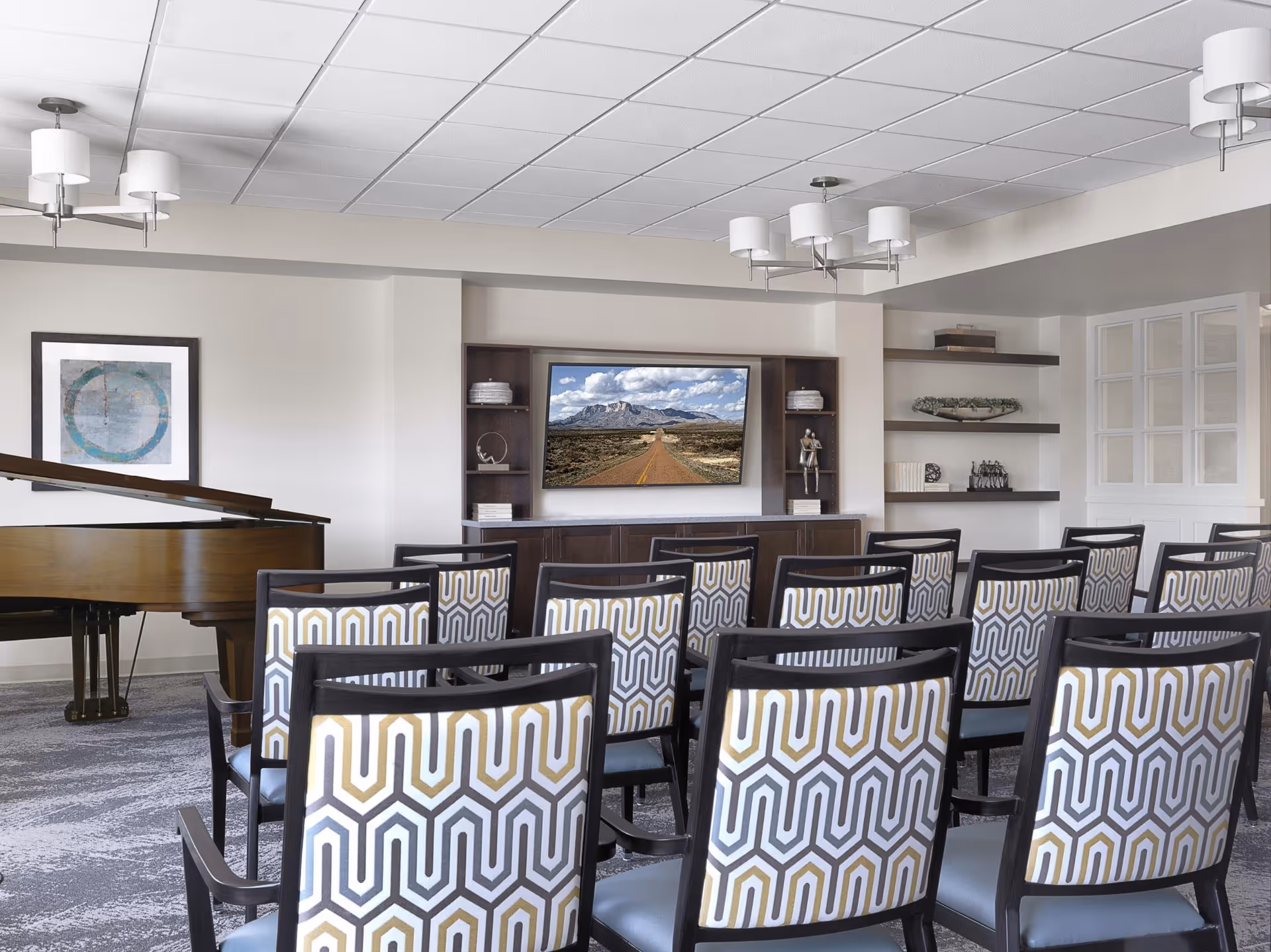 A well-lit room with rows of patterned chairs facing a large flat-screen TV mounted on a wall unit with shelves. To the left, there is a grand piano and a framed abstract artwork on the wall. The room has a modern ceiling with multiple light fixtures and a carpeted floor.