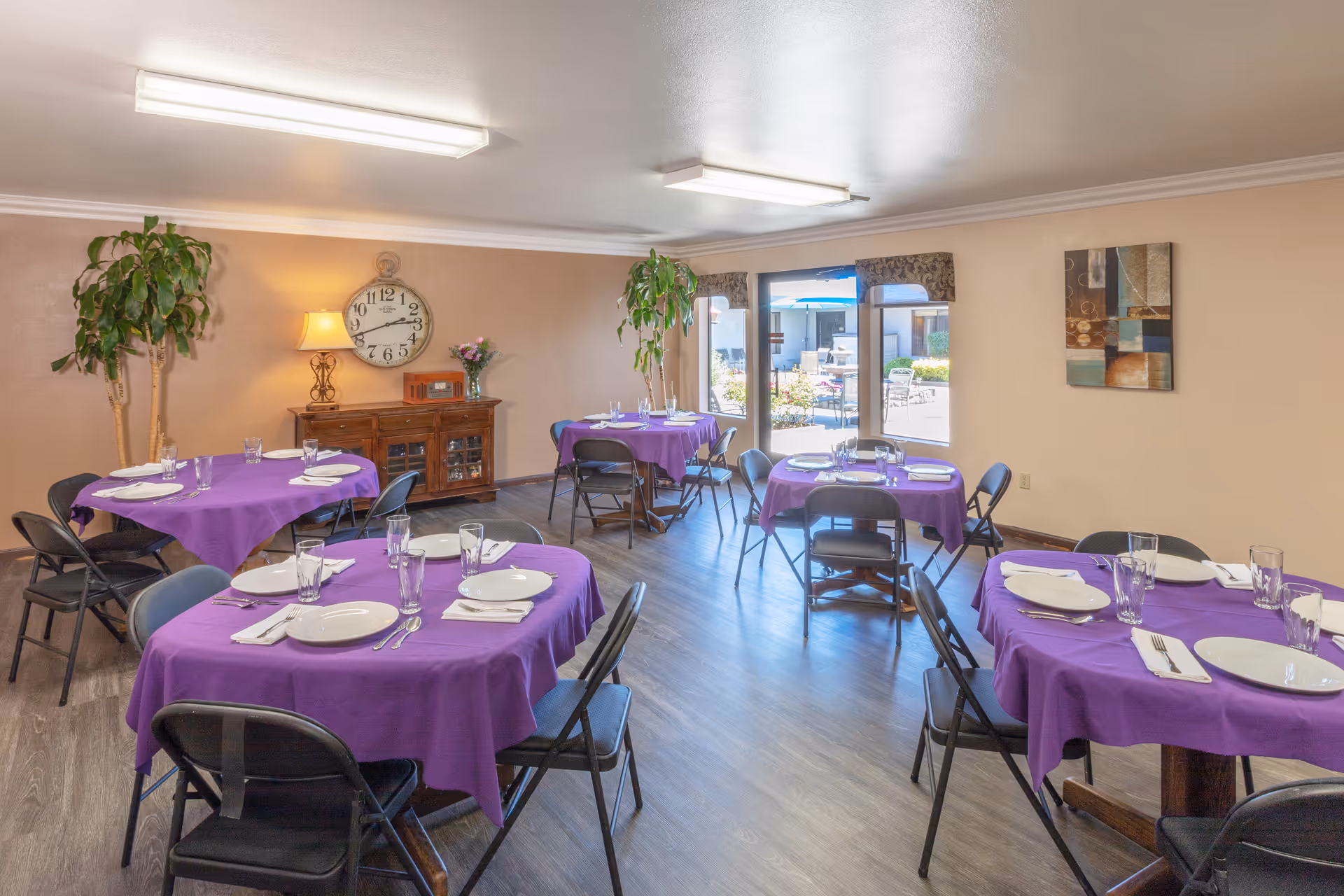 A dining room with several round tables covered with purple tablecloths, each set with white plates, glasses, napkins, and silverware. Black folding chairs surround the tables. The room has wood flooring, beige walls, and large windows letting in natural light. There are two tall green plants, a wooden sideboard with a lamp, clock, and flowers, and a piece of abstract wall art. Outside the windows, there is a patio area with tables and umbrellas.