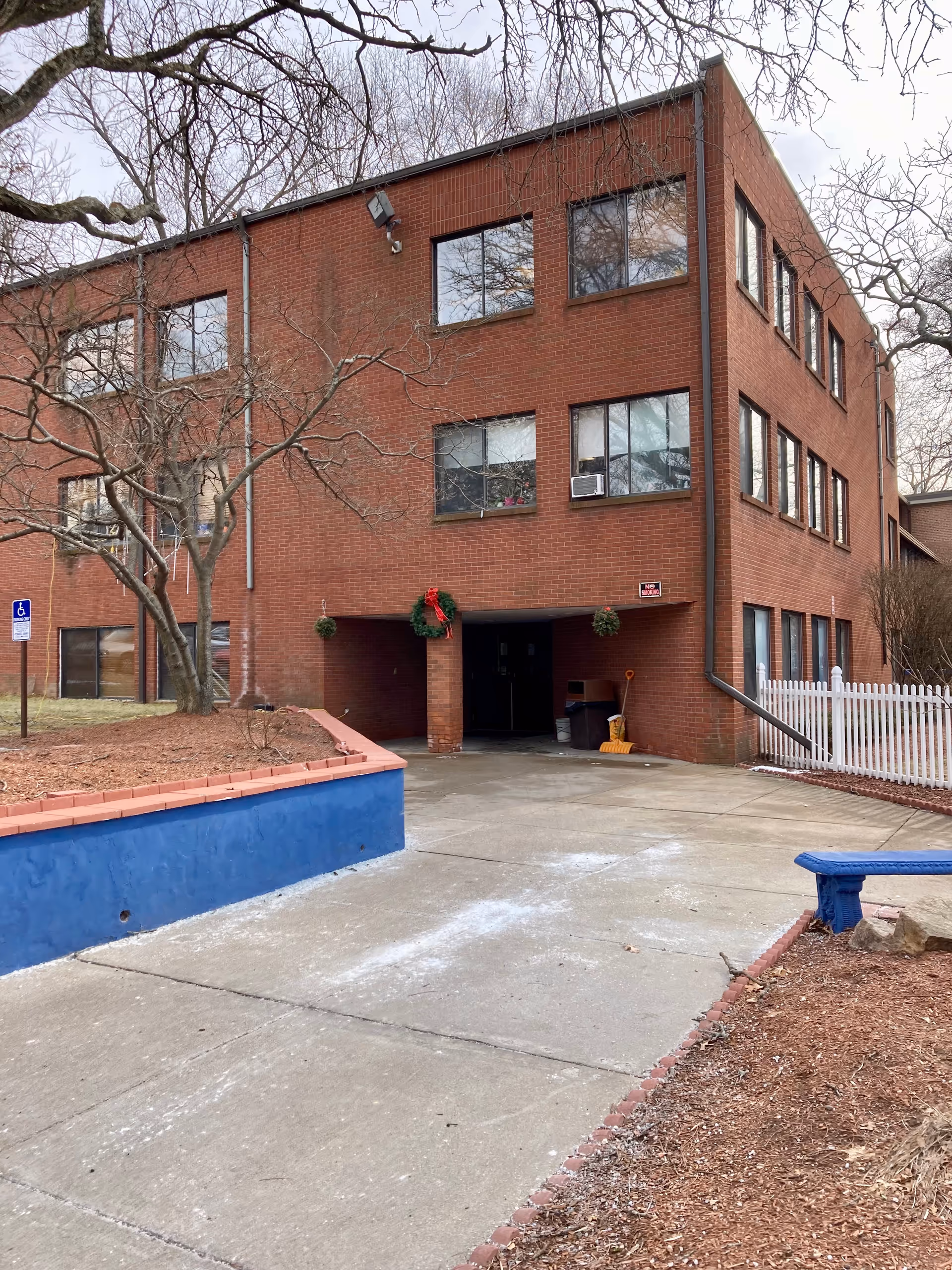 Three-story red-brick building with a covered entrance, concrete driveway, wreath decorations, and bare trees.