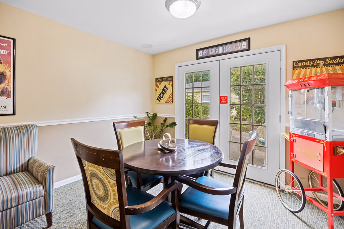 A cozy game room with a round wooden table surrounded by four chairs, each with different patterned upholstery. To the left is a striped armchair, and to the right is a vintage-style red popcorn machine. French doors with glass panes lead outside, and the walls are decorated with vintage-style signs including one that says 'GAME ROOM'.
