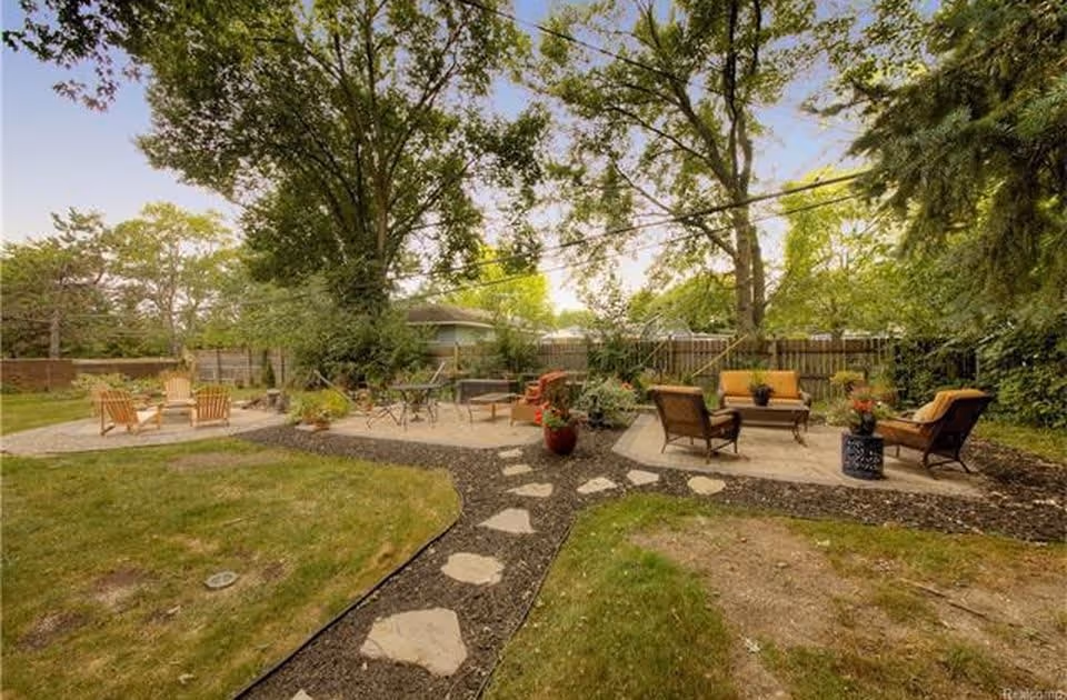 Outdoor garden area with multiple seating arrangements including wooden Adirondack chairs around a fire pit, metal chairs and tables, and cushioned patio chairs with a small table. The area is surrounded by trees and a wooden fence, with stone pathways connecting the seating areas.