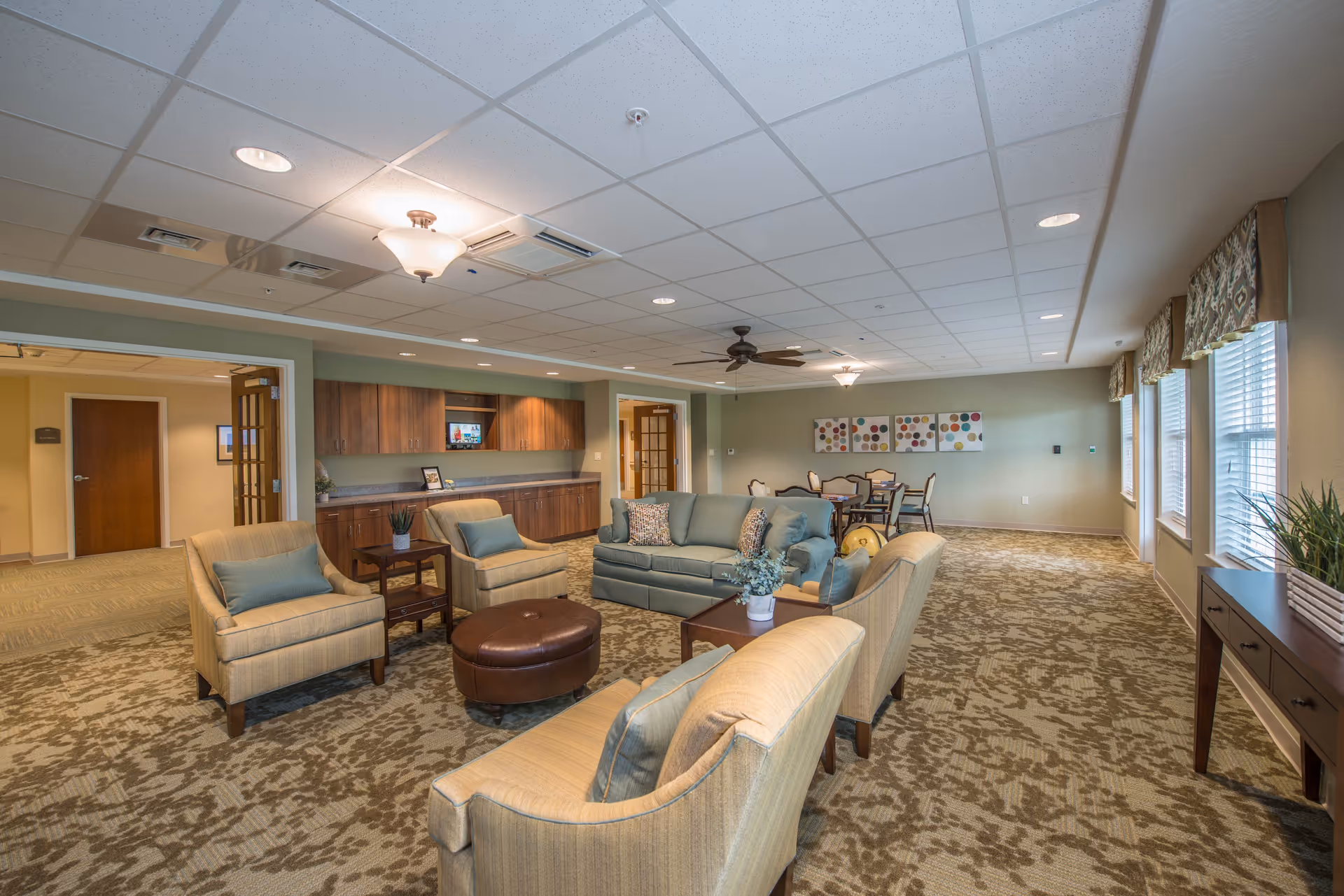 A spacious and well-lit living room area in an assisted living facility featuring a seating arrangement with a sofa, armchairs, a round ottoman, and side tables. The room has carpeted floors with a patterned design, a ceiling fan, recessed lighting, and large windows with valances letting in natural light. There is a wooden cabinet with a small TV mounted above it and a dining table with chairs in the background.
