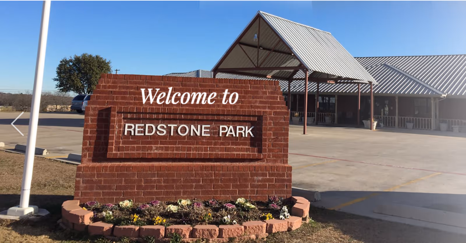 Brick entrance sign reading "Welcome to REDSTONE PARK" in front of a metal-roofed facility entrance and parking area.
