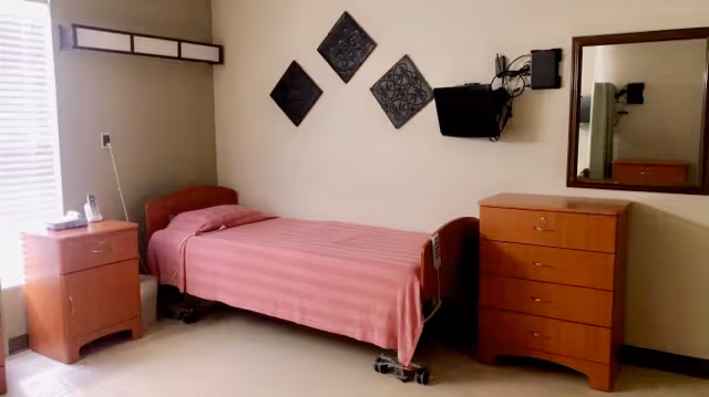 A simple nursing home bedroom with a single bed covered in a pink striped bedspread. The room includes a wooden nightstand with a telephone and a wooden dresser with a mirror above it. There is a small wall-mounted TV and decorative wall art above the bed. A window with blinds is partially visible on the left side.