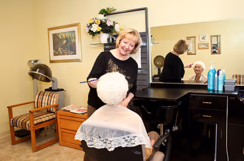 A hairdresser styling the hair of an elderly woman seated in a salon chair in a cozy room with a large mirror, a wooden chair with striped cushions, a side table with magazines, and a flower arrangement on a shelf.