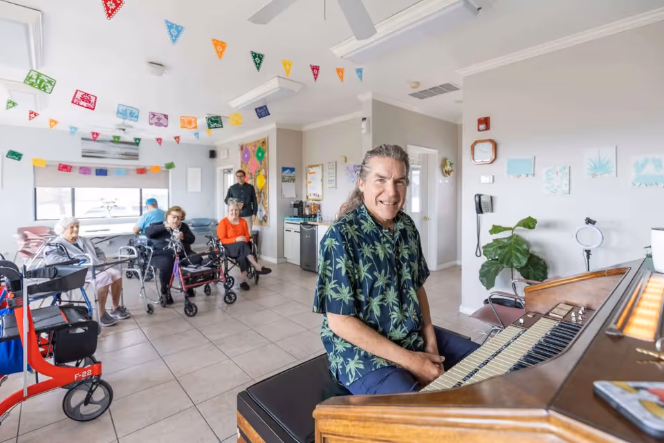 A man wearing a dark shirt with green palm tree prints is sitting at a piano and smiling. In the background, several elderly people are seated in a bright room decorated with colorful triangular paper banners hanging from the ceiling. Some of the elderly people are using walkers and appear to be enjoying the music or socializing.