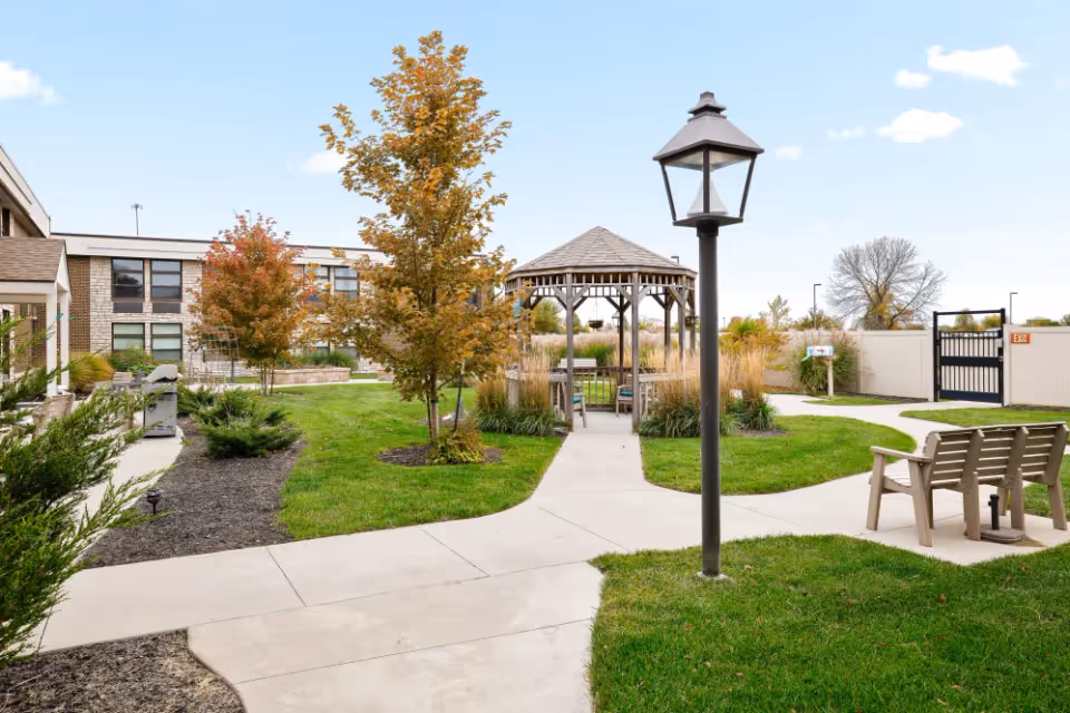 Outdoor courtyard area at Copper Creek Assisted Living & Memory Care featuring a paved walkway, a wooden gazebo with seating, a lamppost, benches, green grass, and trees with autumn foliage under a partly cloudy sky.