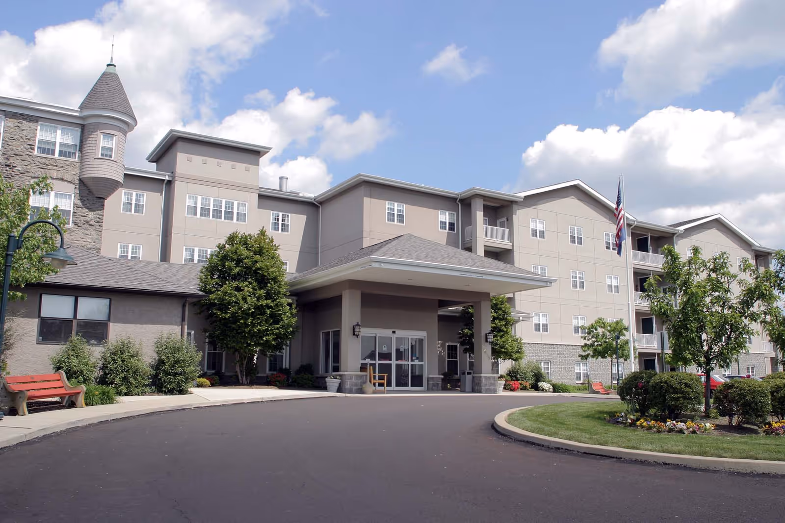 Exterior view of a multi-story senior living facility with a covered entrance, landscaped greenery, benches, and an American flag on a flagpole under a partly cloudy sky.