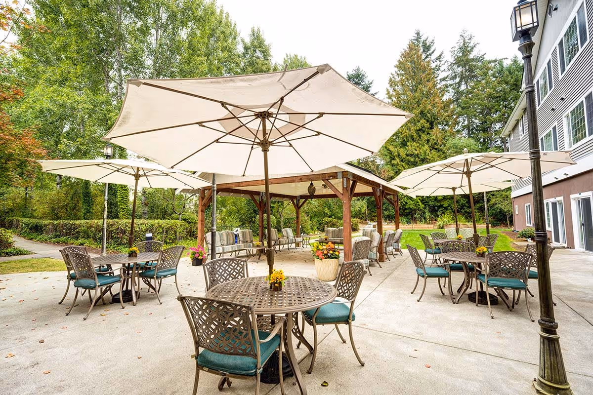 Outdoor patio area with multiple round metal tables and chairs with teal cushions, shaded by large white umbrellas. There is a wooden pergola with additional seating in the background, surrounded by greenery and trees. The side of a building and a lamppost are visible on the right side.