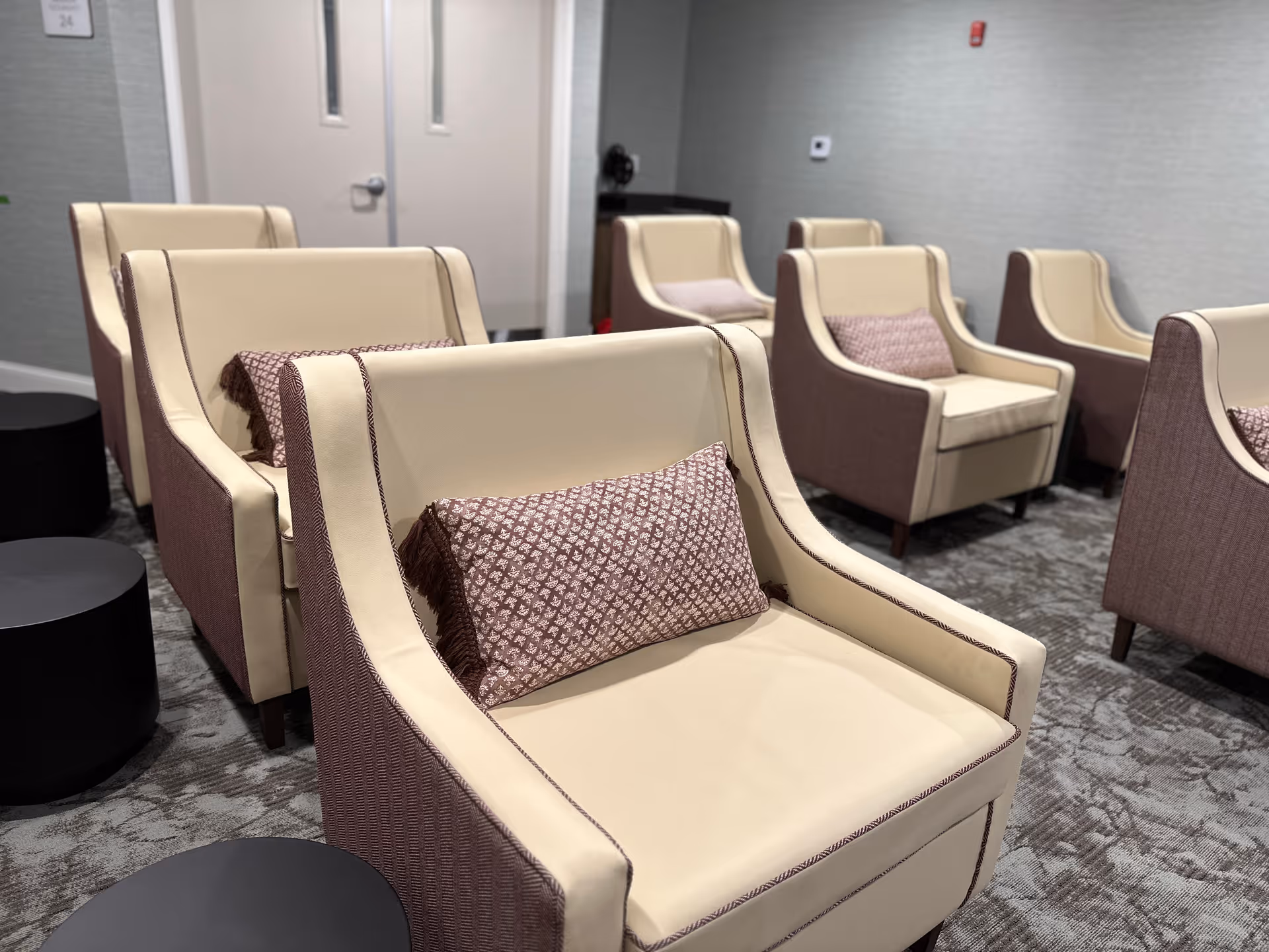 A room with multiple beige and brown armchairs arranged in rows, each with a patterned cushion. The room has a carpeted floor and light gray walls with a closed double door in the background.