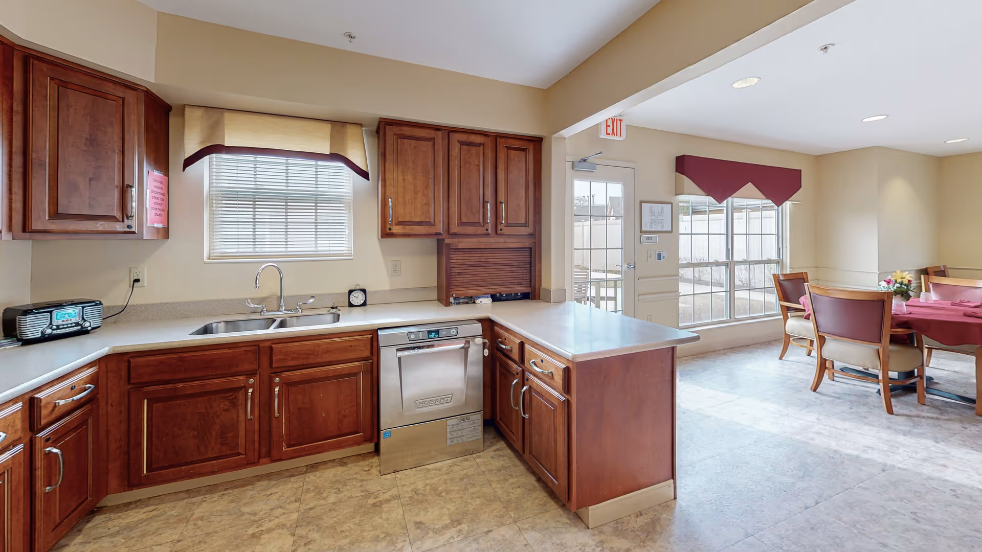 A kitchen area with wooden cabinets, a stainless steel dishwasher, a double sink under a window with blinds and a beige valance. To the right, there is a dining area with a table covered with a red tablecloth and several wooden chairs. Large windows with red valances let in natural light. The floor is tiled and there is an exit door in the background.