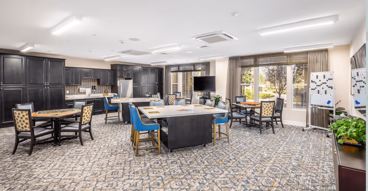 A spacious, well-lit common area in a senior living facility featuring multiple tables and chairs arranged for group activities or dining. The room includes a large kitchen area with dark cabinetry, stainless steel appliances, and a central island. Large windows with blinds allow natural light to fill the space, and a TV is mounted near the windows. The carpet has a patterned design, and there are plants and a whiteboard in the room.